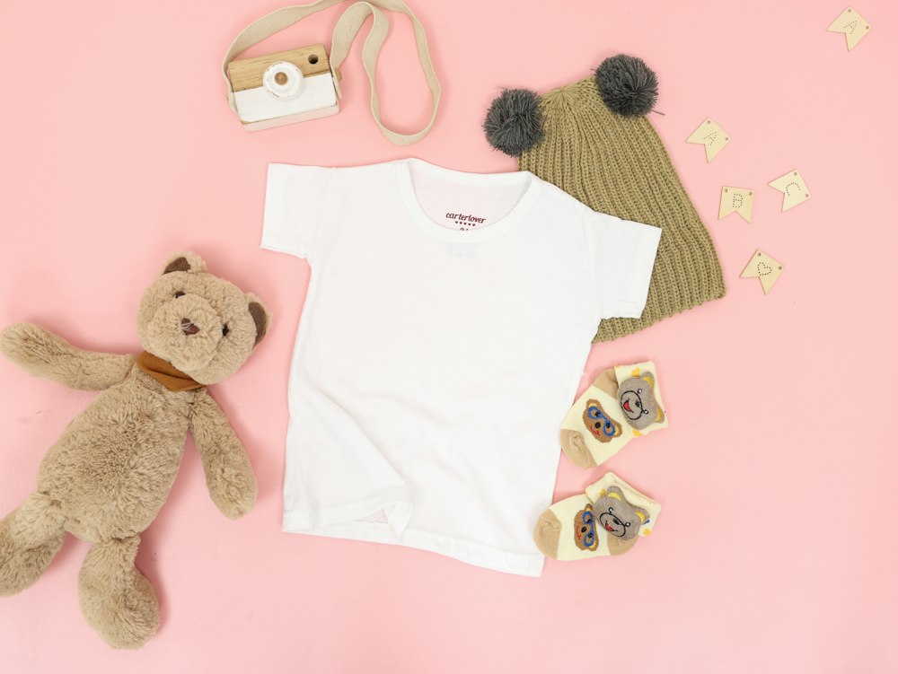 White cotton baby t-shirt with a knitted pom-pom beanie and bear-face socks laid out on a pink background